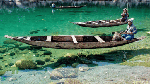 Two wooden boats with people on the clear emerald waters of Dawki river in Meghalaya, with smooth stones visible through the transparent water below.