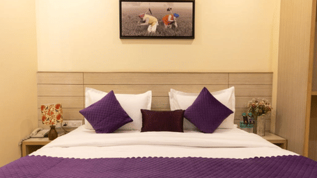 A hotel room, featuring a bed with white linens and a purple runner, a small framed painting above the bed, and a small statue on the nightstand | Perfectstayz Value Sagar