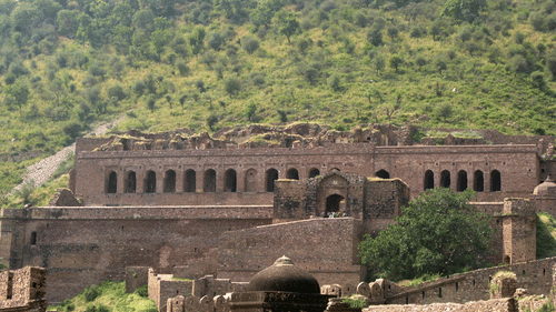 The weathered stone ruins of Bhangarh Fort stand against a backdrop of a lush green hillside under the daylight.