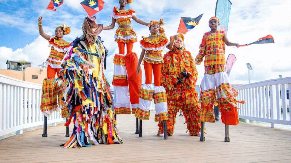 Stilt walkers in patterned, festive attire proudly display Antigua and Barbuda flags outdoors.