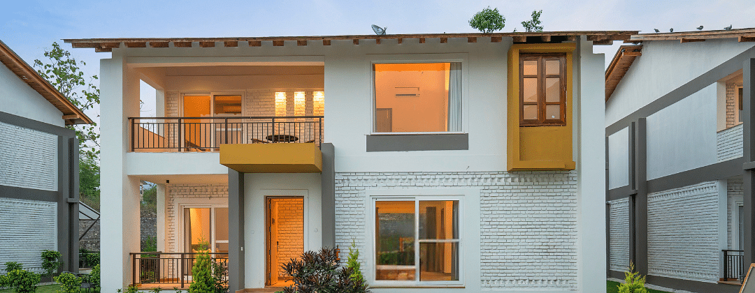 A two-story villa with a balcony, surrounded by lush greenery and a clear blue sky.