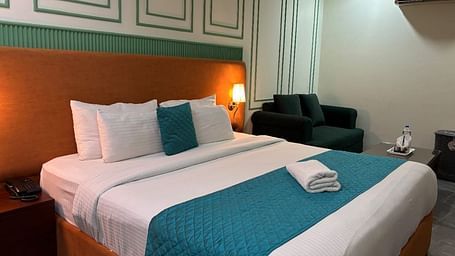 A well-lit room with a double bed dressed in white and blue, and a dark armchair - Asian Suites IFFCO Metro, Gurgaon