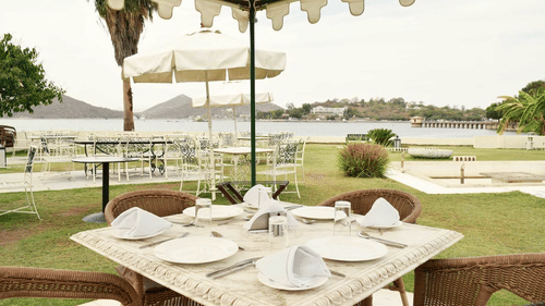 Outdoor dining setup at Ram Pratap Palace, Udaipur with a marble table, chairs, plates, and napkins under a canopy, overlooking the lake and distant hills.