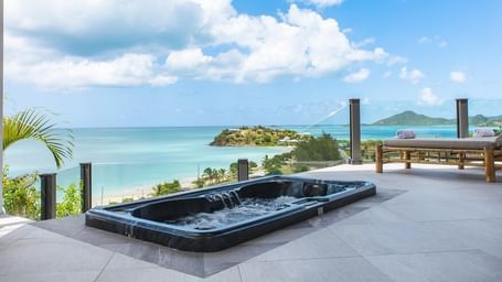  A stunning outdoor jacuzzi at Tamarind Hills Resort and Villas - Antigua, offering panoramic views of a turquoise ocean and a clear blue sky with scattered clouds. The jacuzzi is black and appears to be built into a light-colored patio.