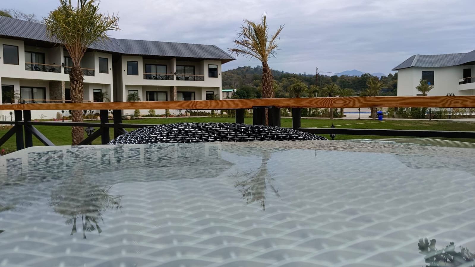 A view over a glass table reflecting a resort with palm trees and mountains in the distance - Shervani Pebbles & Pines, Corbett