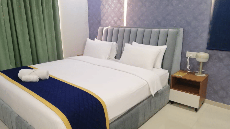 A contemporary hotel bedroom with a large double bed, blue quilted headboard, and a matching decorative bed runner at Apartel by Aarin, Oragadam.