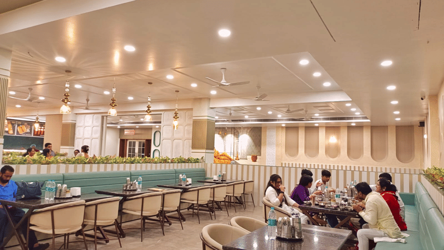 A wide interior shot of the Maharani Ki Rassoii, showing a spacious, well-lit dining area with light green booth seating, wooden tables, and several patrons dining.