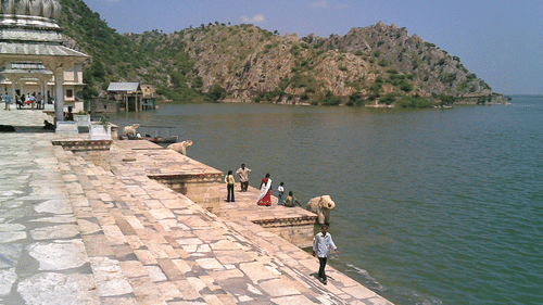 View of a lake with rocky hills and a clear sky in the background and people along the paved shoreline near a domed structure.