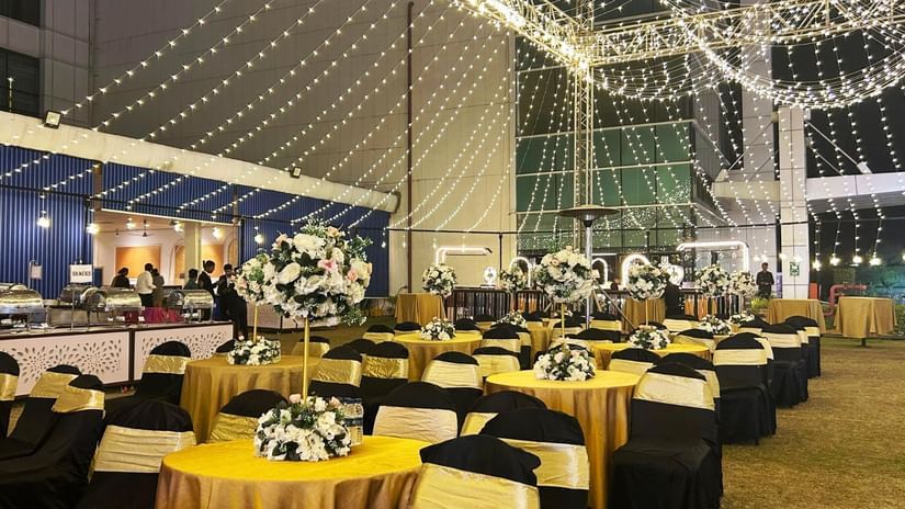 An outdoor nighttime event setup with many round tables and chairs, illuminated by strands of overhead string lights, with a buffet area visible on the left.
