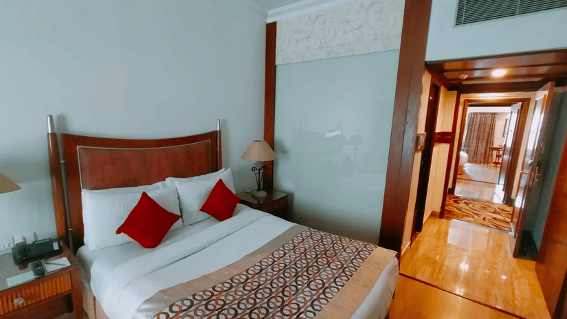 A bedroom at Hotel Hindusthan International, Kolkata, featuring a comfortable bed, bedside tables, and a corridor leading into a spacious suite area.