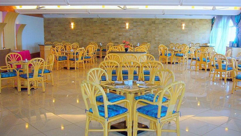 Interior of Flavors Restaurant with round tables and wooden chairs with blue cushions, under warm lighting at Kohinoor Samudra Beach Resort, Ratnagiri