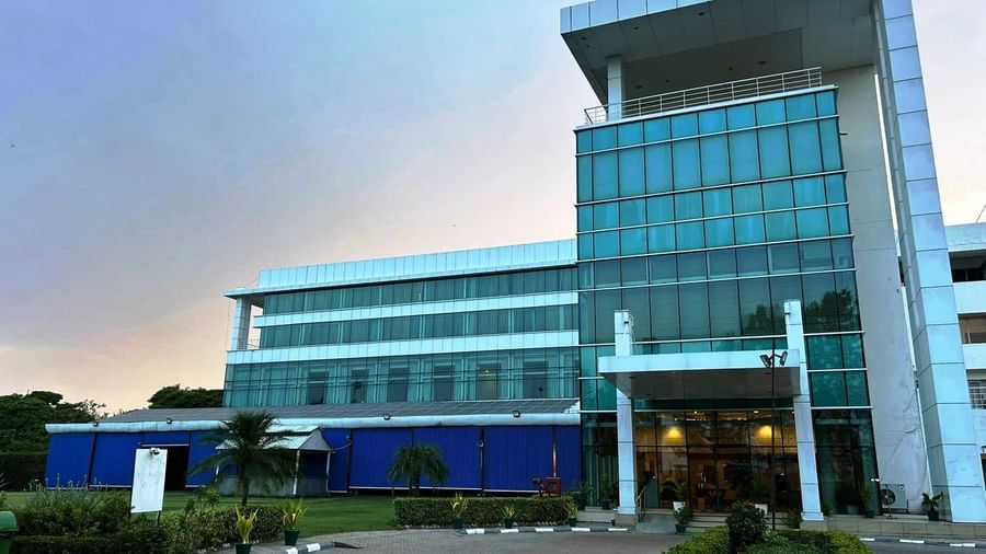 An exterior view of the modern, multi-storey Amer Greens hotel building, featuring a prominent glass facade and a manicured driveway and lawn.