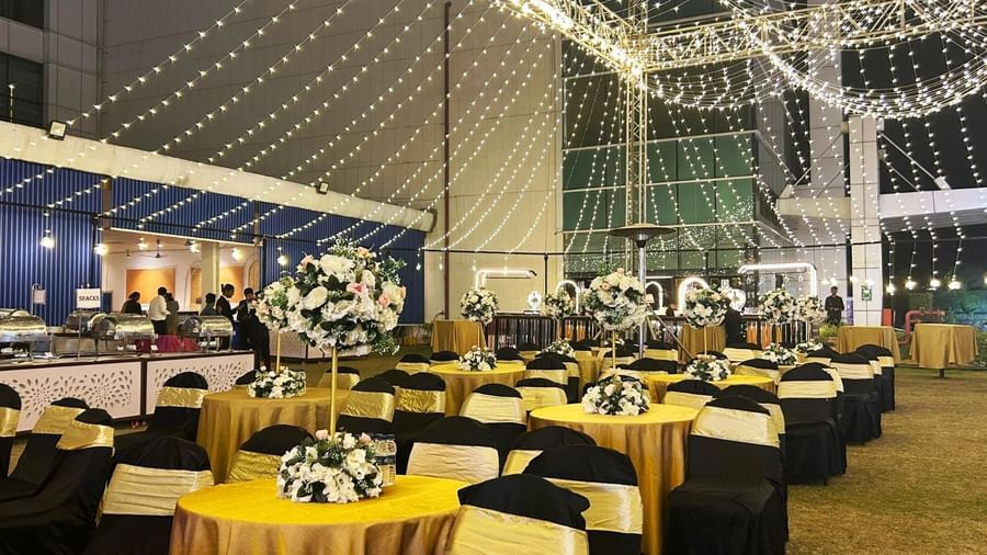 An outdoor nighttime event setup with many round tables and chairs, illuminated by strands of overhead string lights, with a buffet area visible on the left.