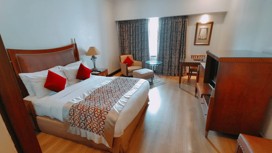 A hotel room at Hotel Hindusthan International, Kolkata, featuring a double bed, study area, bright lighting, and wooden flooring.