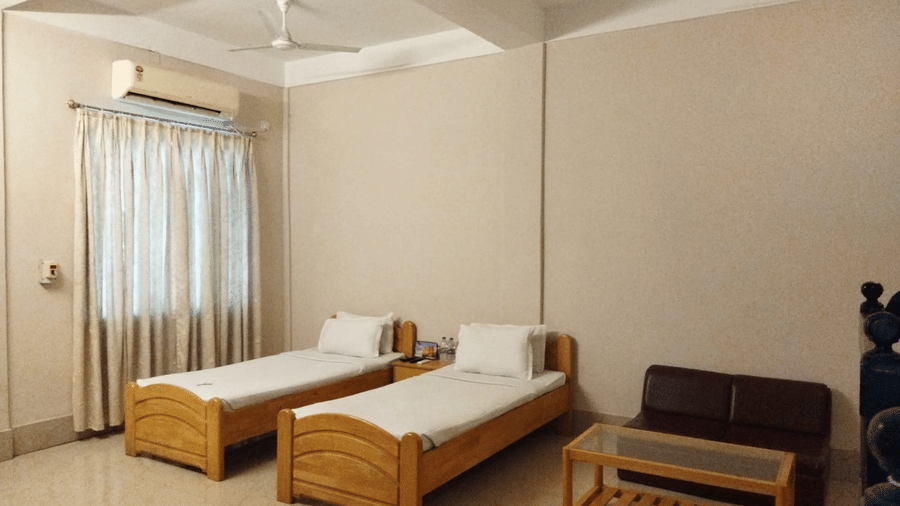 Interiors of a room at Lake Side Resort Neermahal featuring 2 wooden beds, an A/C unit, a small wooden table and tiled floors.