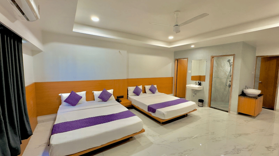 Interior of a clean, well-lit room at Pushpak Resort, Shirdi, with double beds and a glass-partitioned area.