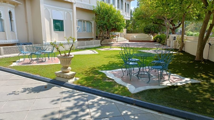 Garden area with green lawn, trees, metal chairs, and tables placed near the building facade at Ram Pratap Palace, Udaipur.