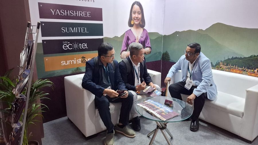 Three men in formal attire seated on a white sofa discussing at the Sumi Yashshree Hotels booth, with branding and a scenic backdrop.
