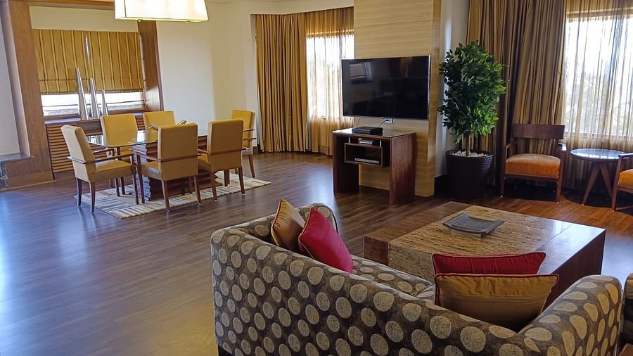 A spacious living area featuring a patterned sofa, coffee table, wall-mounted TV, and a dining table with upholstered chairs near large windows - The Retreat Hotel and Convention Centre