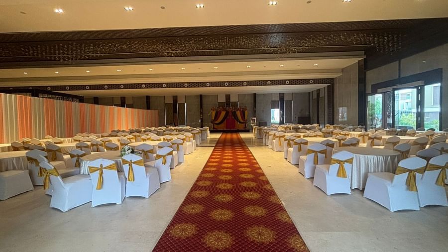 A well-lit banquet hall with rows of white chairs and a red carpet, leading to a stage at the far end.