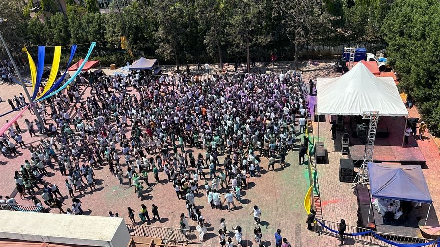Aerial view of a large crowd gathered outdoors, colorful flags and tents visible, sunny day.