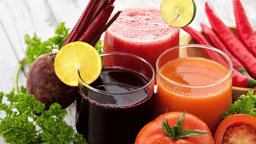 3 glasses of assorted vegetable juices surrounded by fresh vegetables, including tomatoes and limes