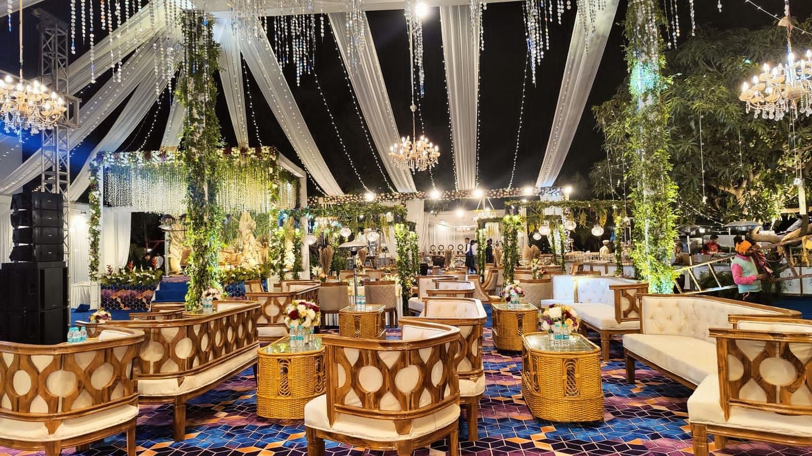A beautifully decorated outdoor wedding venue with chandeliers, draped fabric, and seating arrangements on a colorful carpet.