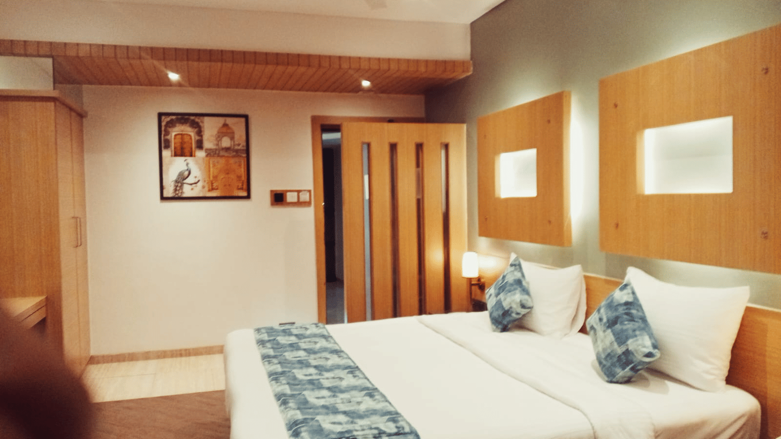 A bedroom space arranged with double beds, a wall-mounted wooden frame, an overhead ceiling fan, and a light-toned wooden wardrobe unit at Hotel Mumbai House, Thane.