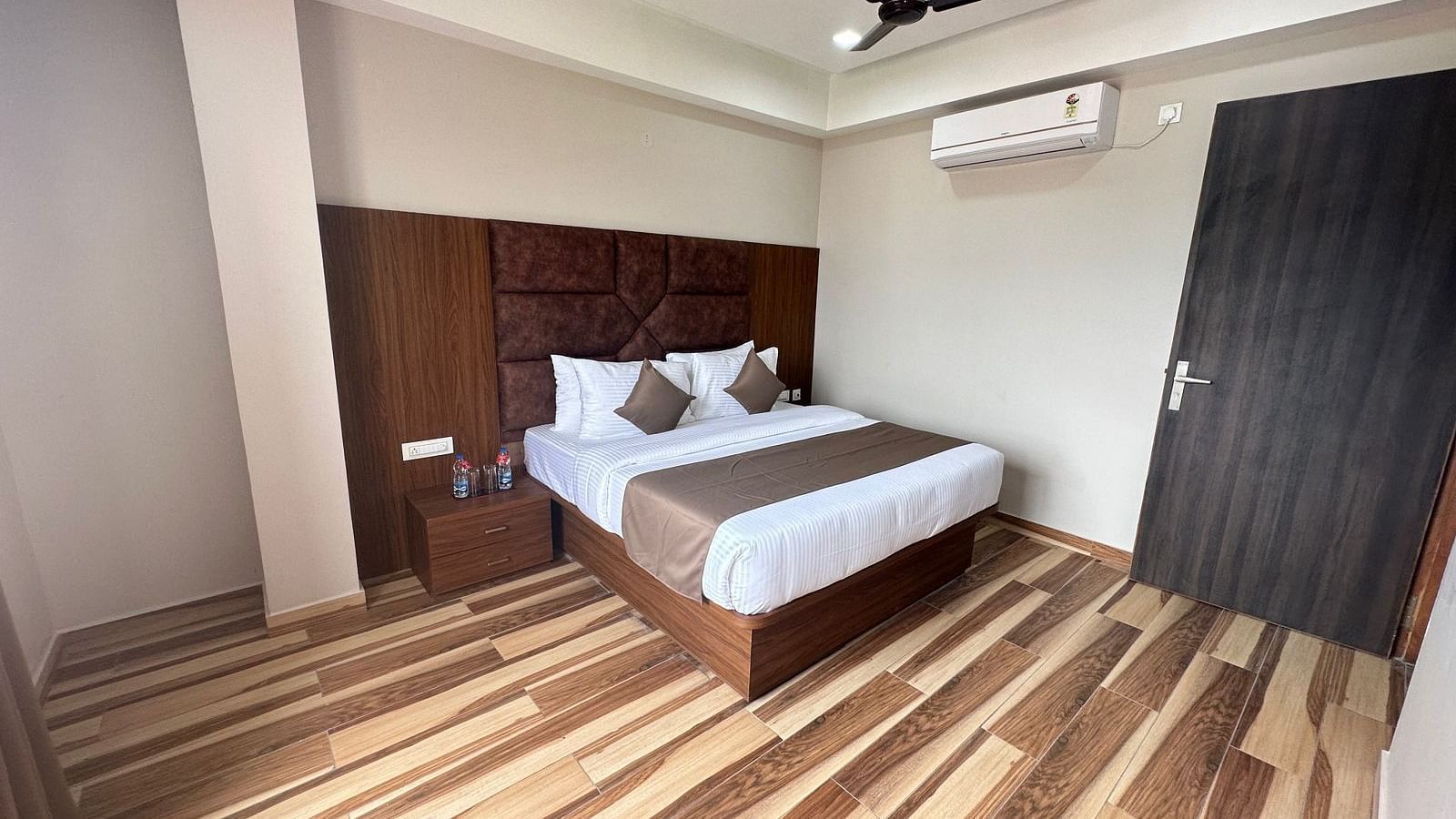 A bedroom includes a double bed with white and beige linens, dark wooden furniture, and a ceiling fan - Kankidham Lords Inn