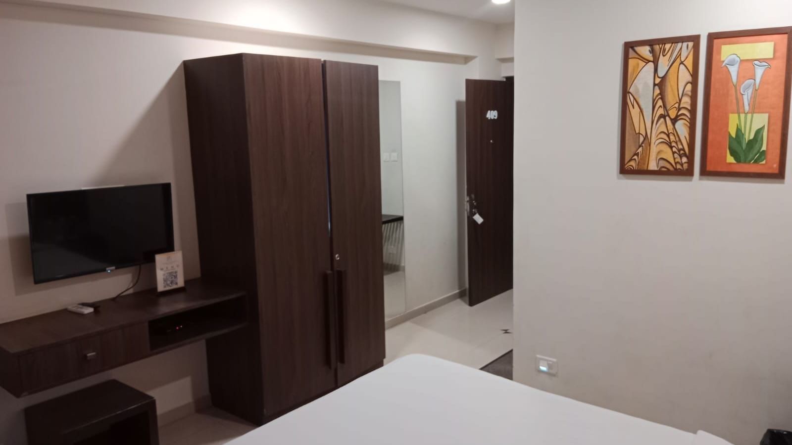 Full view shot of the Deluxe Room at Pravasa Ahmedabad featuring a wall mounted television beside a wooden wardrobe placed opposite to a double bed.