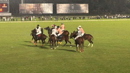 Polo players on horseback contesting for the ball on the field in front of Jaipur Polo Season billboards during a night match.