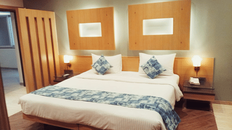 A king size bed with a white duvet, a patterned bed runner, 2 wooden nightstands with lamps, and 2 rectangular wall design panels at Hotel Mumbai House, Thane.