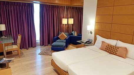 Image of twin beds in the executive twin room at our hotel featuring a flat screen television, bed side table, mirror, violet curtains, and other furniture. | Mirage Hotel