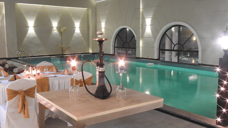 Polo Hotel, Agartala poolside dining area with indoor pool, arched windows, hookah on table, draped chairs with gold sashes and wall sconces.