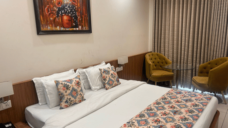 A Premium Room at the Prince ZENQ Xpress Resortt Rohtak featuring a large bed with a floral runner, decorative pillows, a painting on the wall, and two velvet armchairs with a glass side table.