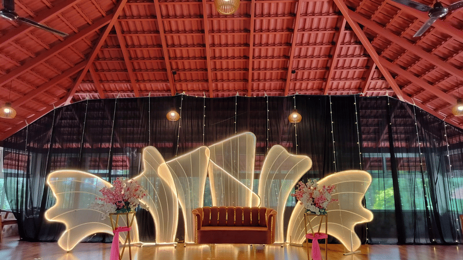 An indoor stage area with a high, exposed truss roof and a large, abstract illuminated white wing-like backdrop at Coorg Orange Blossom Resort and Spa.