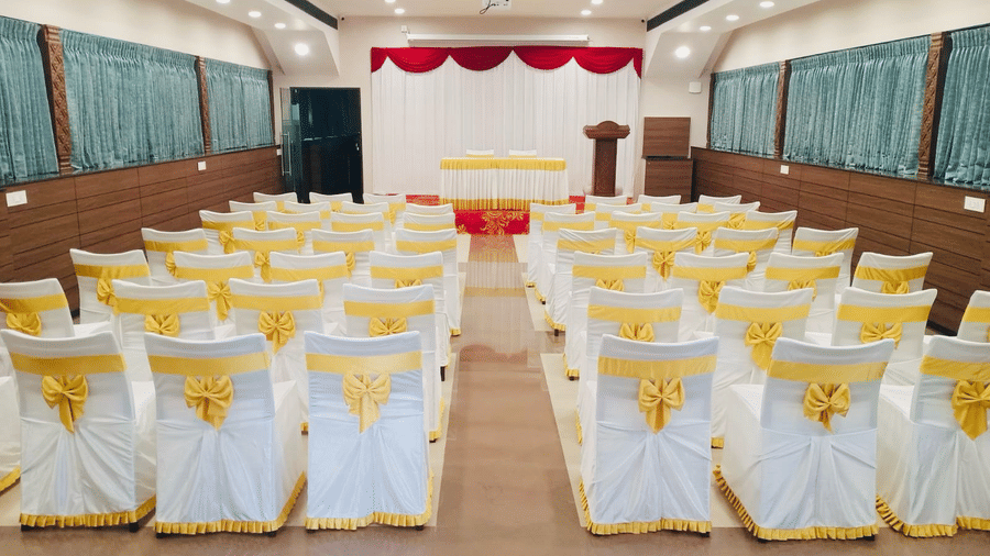 Centered aisle view of Gangai Hall showing white covered chairs with yellow ribbons and a stage in the background.