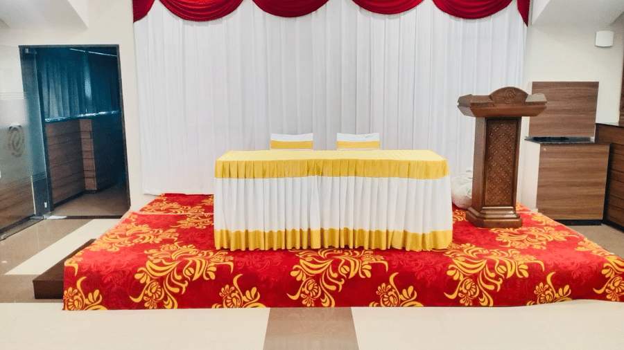Raised stage area inside Gangai Hall featuring a wooden podium, head table with yellow cloth, and white backdrop curtains at Hotel Grand Sabarees Madurai.