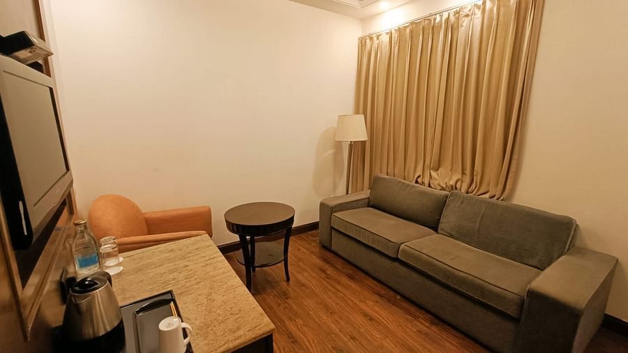 The suite's separate living room features a large sofa, wall-mounted television with a wooden table, lamp, and a single seating cushion at Inde Signature Tower, Gurgaon