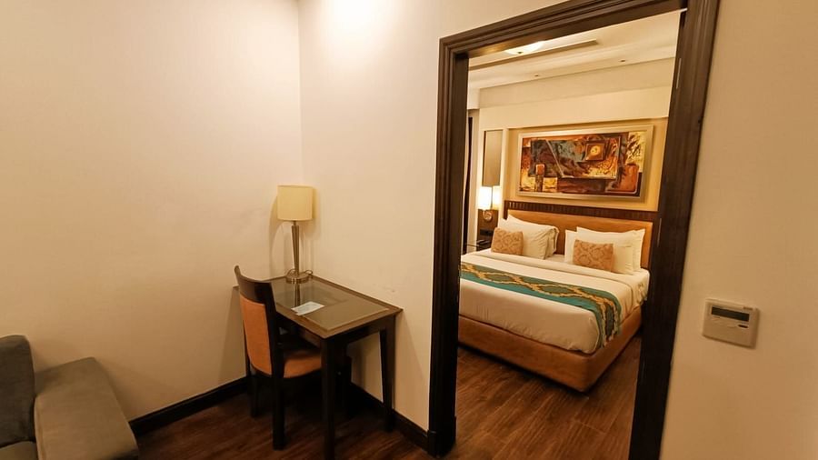 Suite view from separate living area with a double bed neatly arranged with plush pillows, bedside tables each topped with a lamp, and a photo frame on the headboard at Inde Signature Tower, Gurgaon