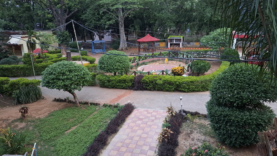 An outdoor garden area at Khairi Resorts has a curved paved path, neatly trimmed bushes, and a central seating spot.
