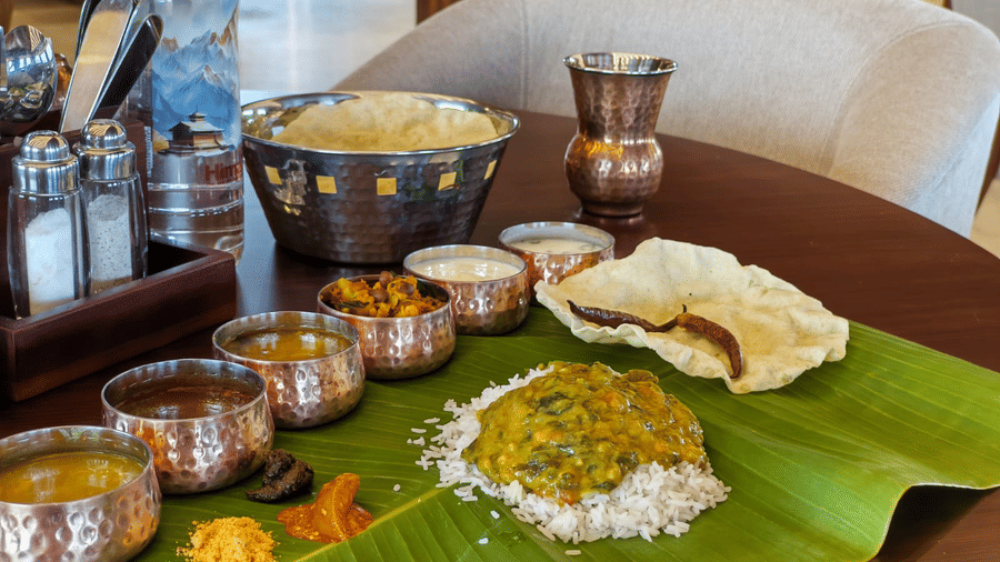 Dining table setup at Ni Central Park, Mysore, showcasing a banana leaf meal with traditional metal serving bowls and drinks in a cozy restaurant setting.