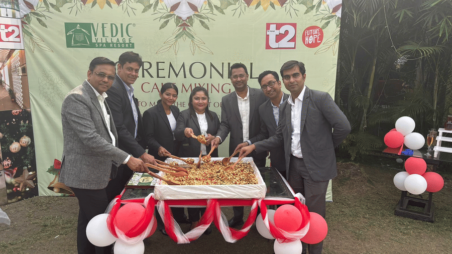 A group of staff members posing together at a ceremonial event at Vedic Village Spa Resort with branded backdrop balloons and a cake displayed on the table in front.