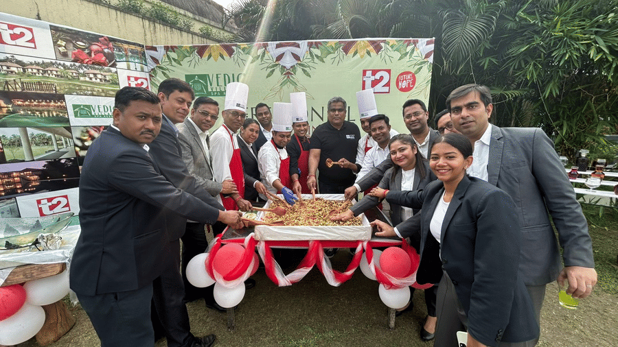 A group of staff members in uniform cutting a cake at a celebration event at Vedic Village Spa Resort with red balloons and event banners visible in the background.