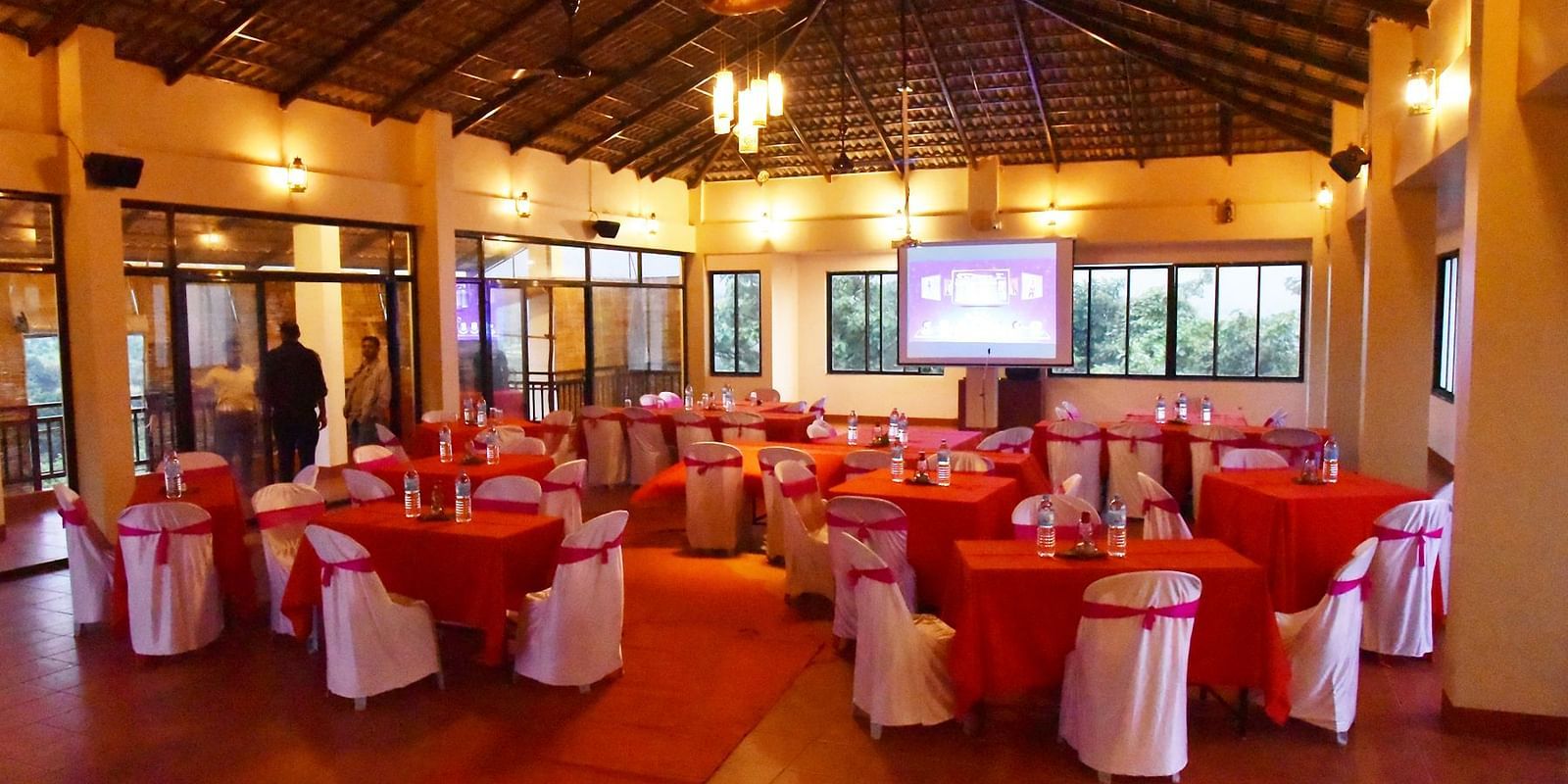 The warmly lit dining hall at Forest Escapes Koyna is set with tables and red-covered chairs.