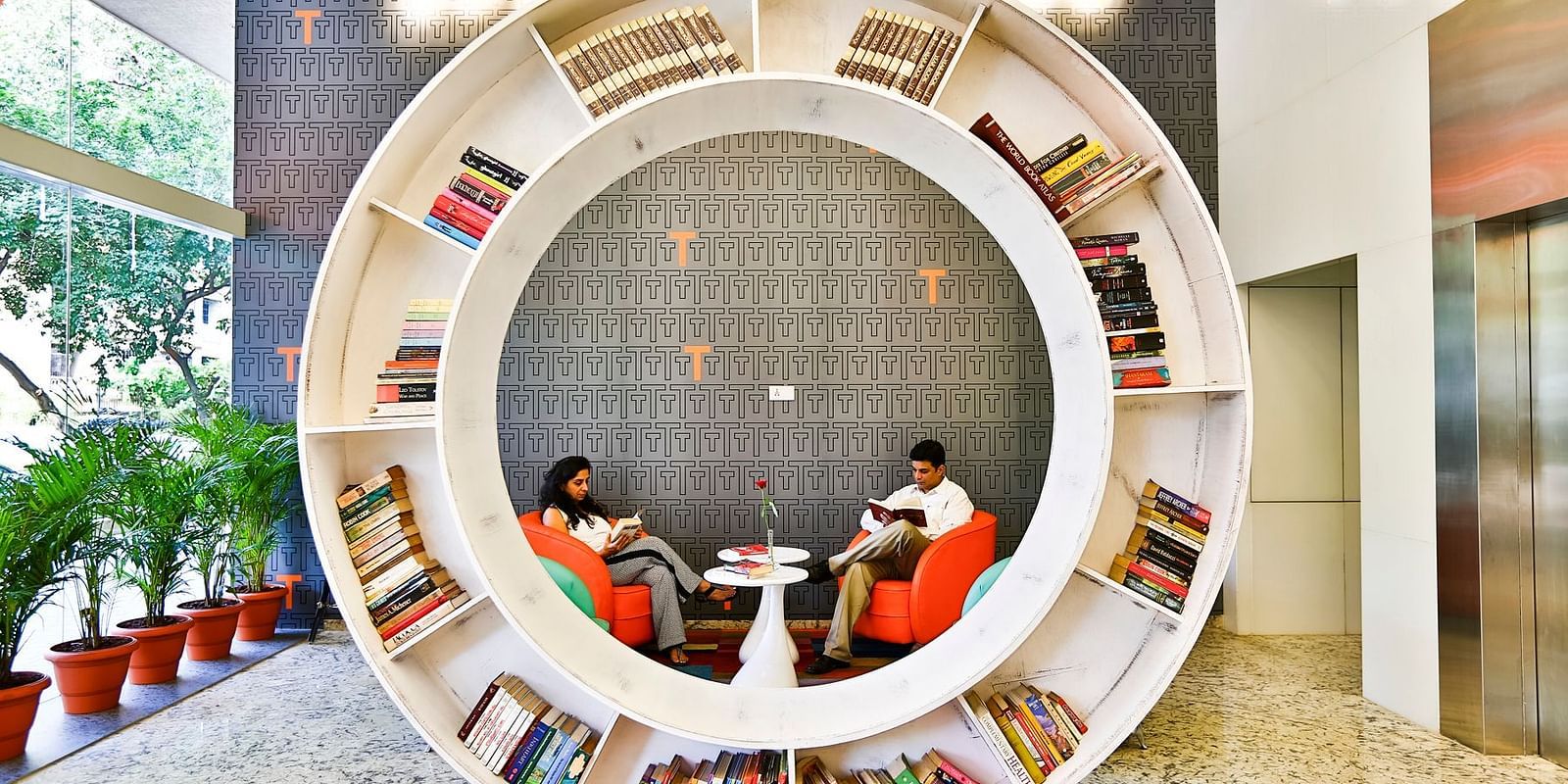 Two people sit in comfortable orange chairs inside a circular bookshelf in the lobby of the T24 Residency hotel in Mumbai, India.