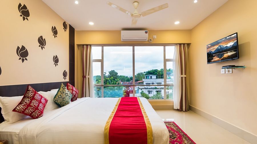 An image of a neatly arranged bed with stylish interior, featuring a wall-mounted TV and a glass window offering a scenic view - Central Courtyard Boutique Hotel, Siliguri