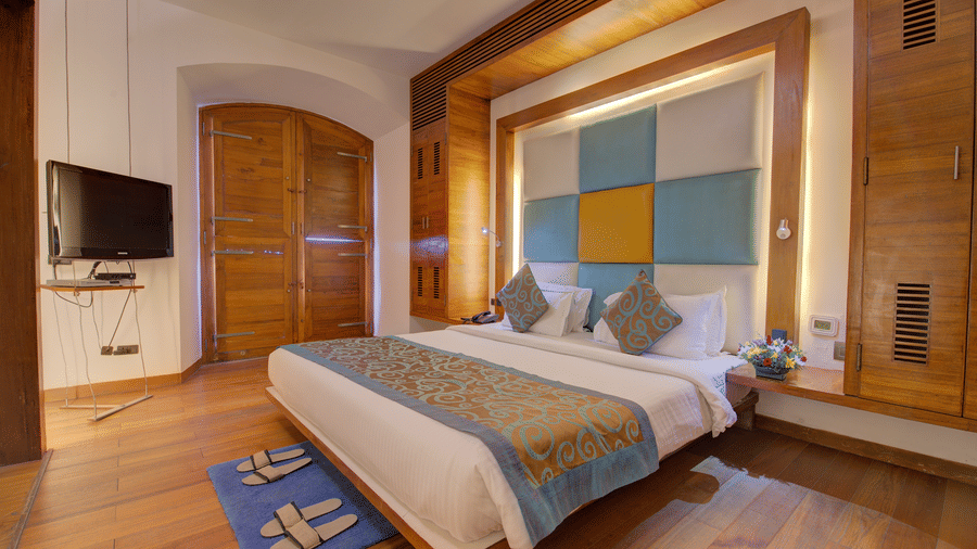 View of a suite bedroom at The Promenade Pondicherry with a king bed, blue and yellow headboard panel, wall-mounted TV, and warm wooden interiors.