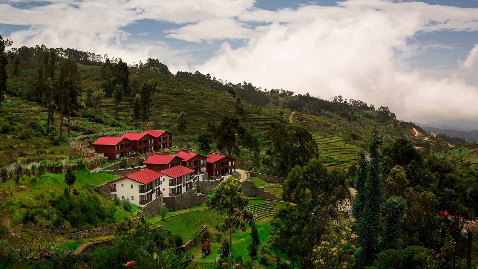 A drone shot of the GReaT Trails Kodaikanal By GRT Hotels property surrounded by lush greenery and trees with clouds hovering over