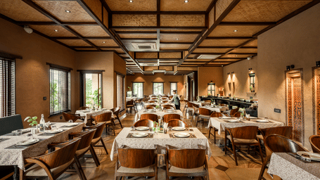 Warmly lit indoor dining area of Baradari at AamaGhati Wildlife Resort with a woven ceiling, rustic walls, and arranged dining tables with cushioned wooden chairs.